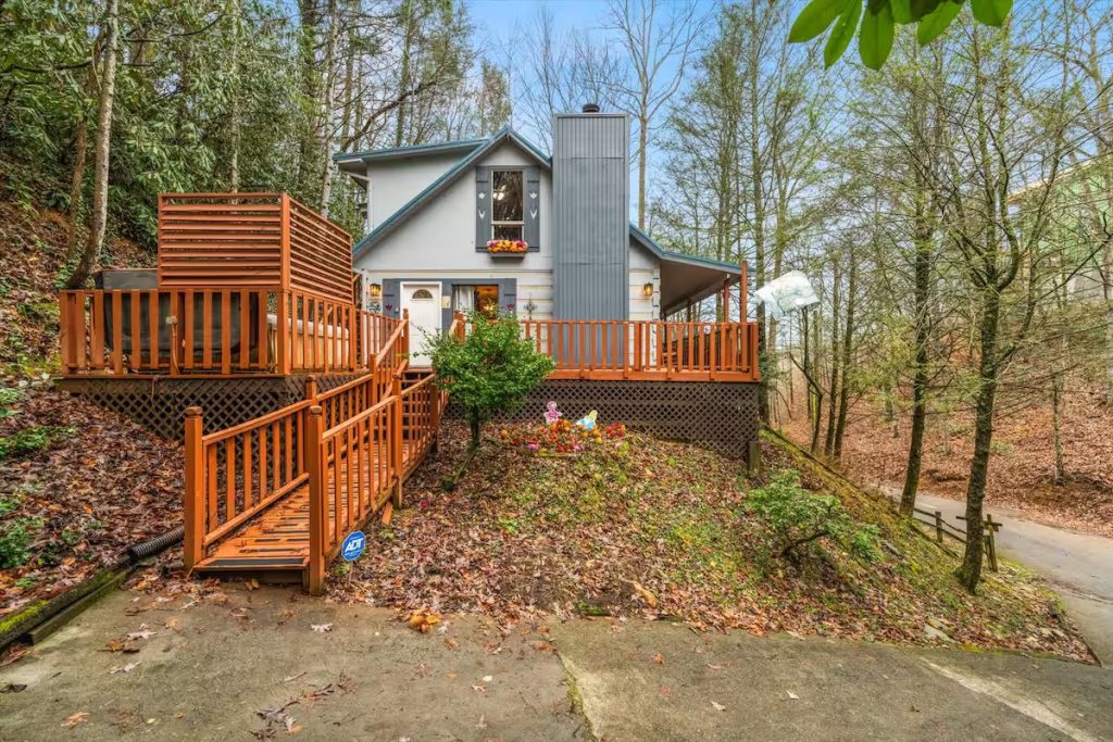 An exterior wide shot of a gray two-story cottage nestled on a wooded hillside. A large wooden wrap-around deck features a privacy screen for a hot tub, a ramp entry, and a small flower bed with whimsical Wonderland-themed figurines.