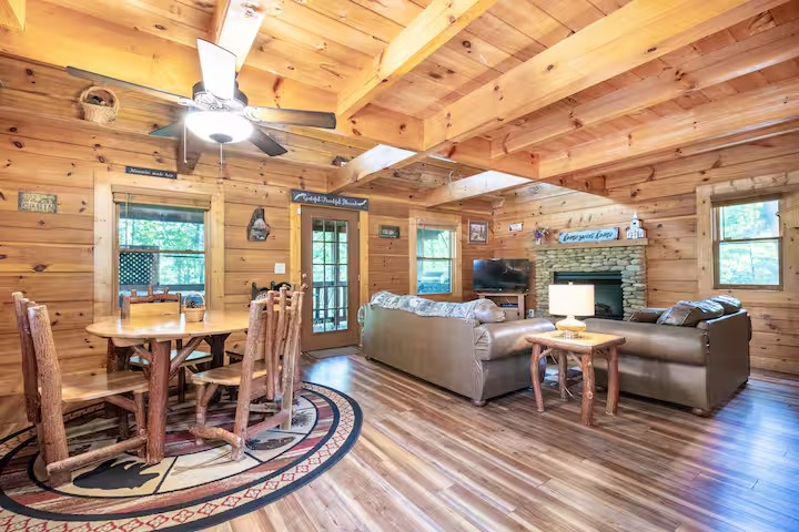 Exterior shot of a two-story wooden log cabin with a green metal roof, nestled in a dense, lush green forest.