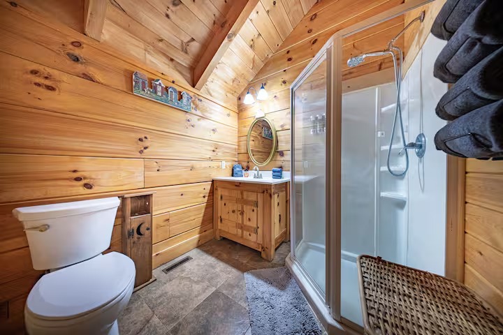 A covered wooden cabin porch featuring a wooden swing with a "Welcome to our Cabin" pillow, overlooking a lush green forest.