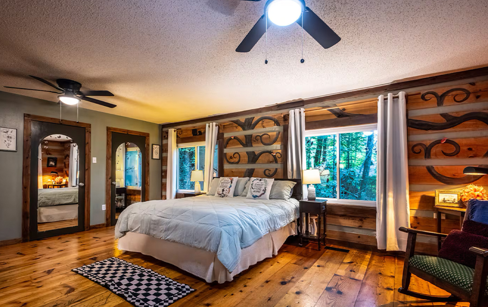 Cozy bedroom inside a Smoky Mountains cabin featuring rustic wooden walls with carved designs, polished hardwood floors, a large comfortable bed, and scenic forest views from the windows.