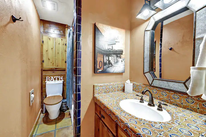Bathroom vanity with a patterned tile countertop, a large metal-framed mirror, and a wood-paneled toilet nook.
