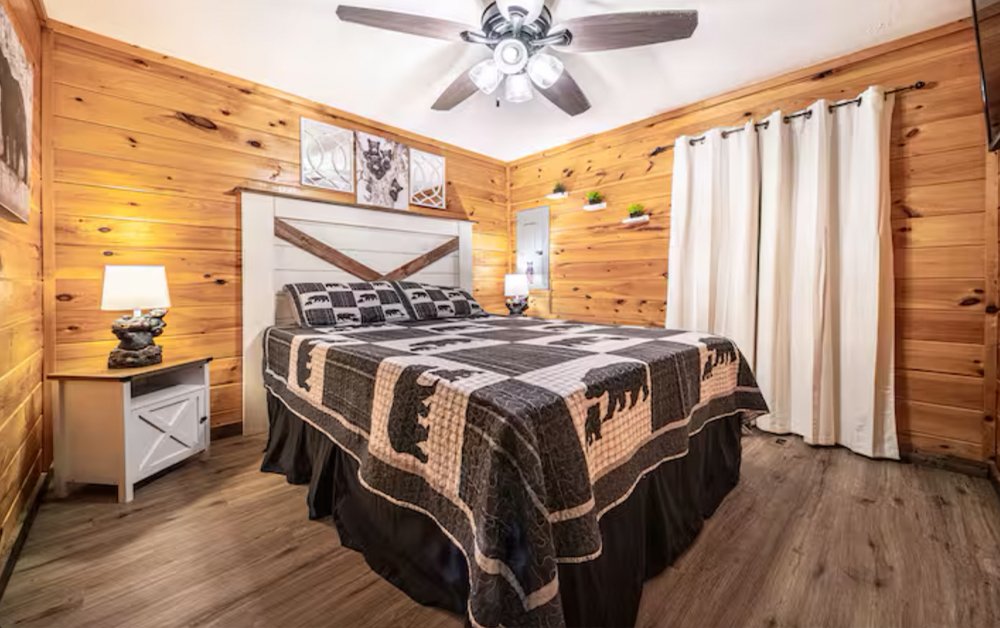 A cozy, premium bedroom in a log cabin with wood-paneled walls, a large bed with a black and white bear-patterned quilt, and modern farmhouse decor, part of the Sevierville Vacation Rentals collection.