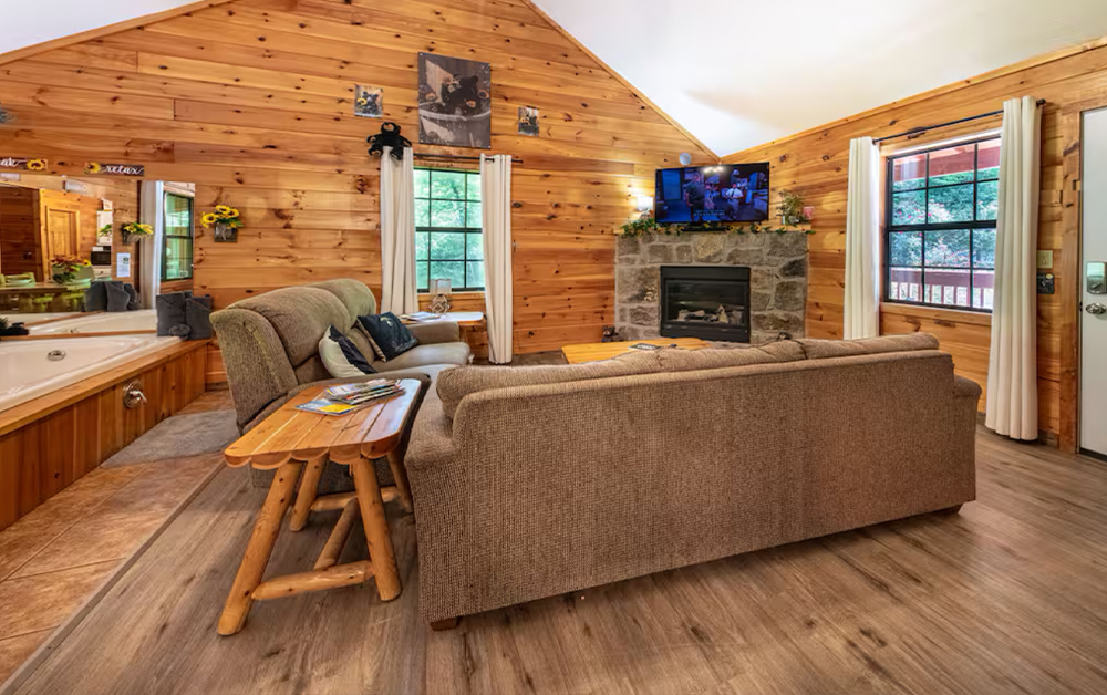 A spacious, open-concept cabin living room featuring wood walls, a stone fireplace, and an integrated indoor jetted tub, a signature of Sevierville Vacation Rentals.