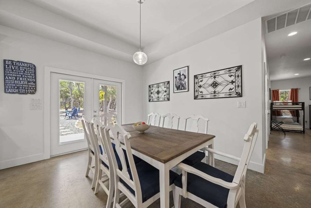 A bright, modern dining area featuring a rustic wooden table with white chairs, polished concrete floors, and a glass door leading to a backyard.