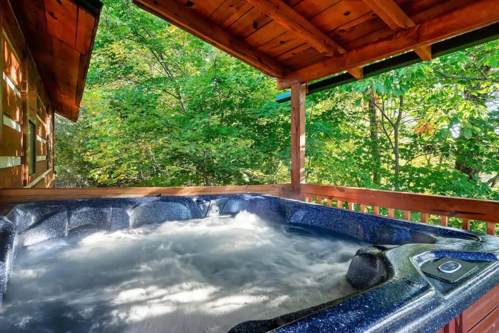 A bubbling hot tub with blue marbled interior sits on a wooden deck of a log cabin, overlooking a dense green forest. The deck is sheltered by a rustic wooden overhang, and the water in the hot tub is steaming and churning.
