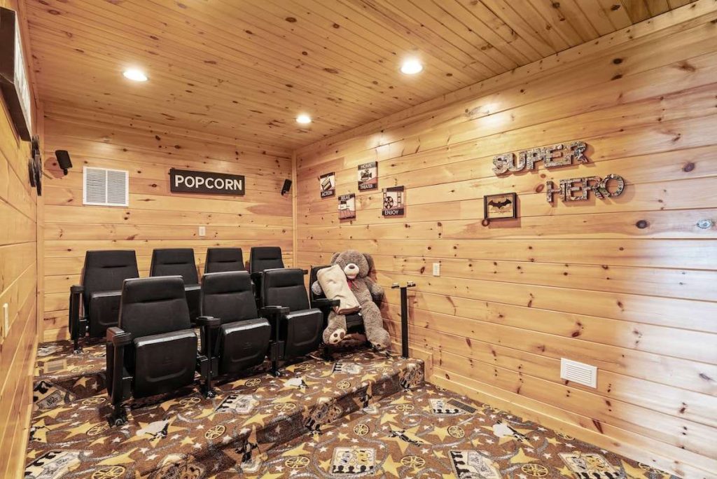 A home theater room inside a log cabin featuring tiered black cinema seating, wood-paneled walls and ceiling, "Popcorn" and "Super Hero" wall decor, and a large teddy bear.