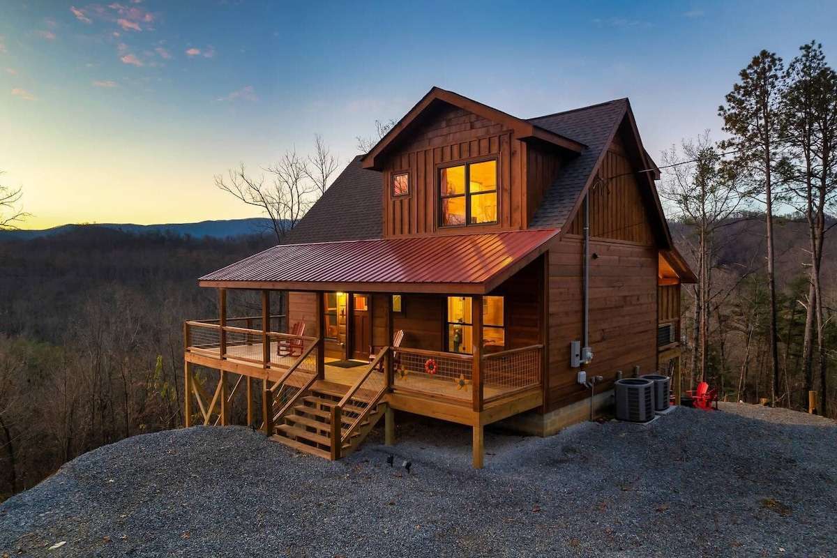 A two-story rustic wooden cabin with a red metal porch roof sits on a gravel lot overlooking a mountain valley at dusk. The cabin features warm interior lighting and a wrap-around wooden deck.