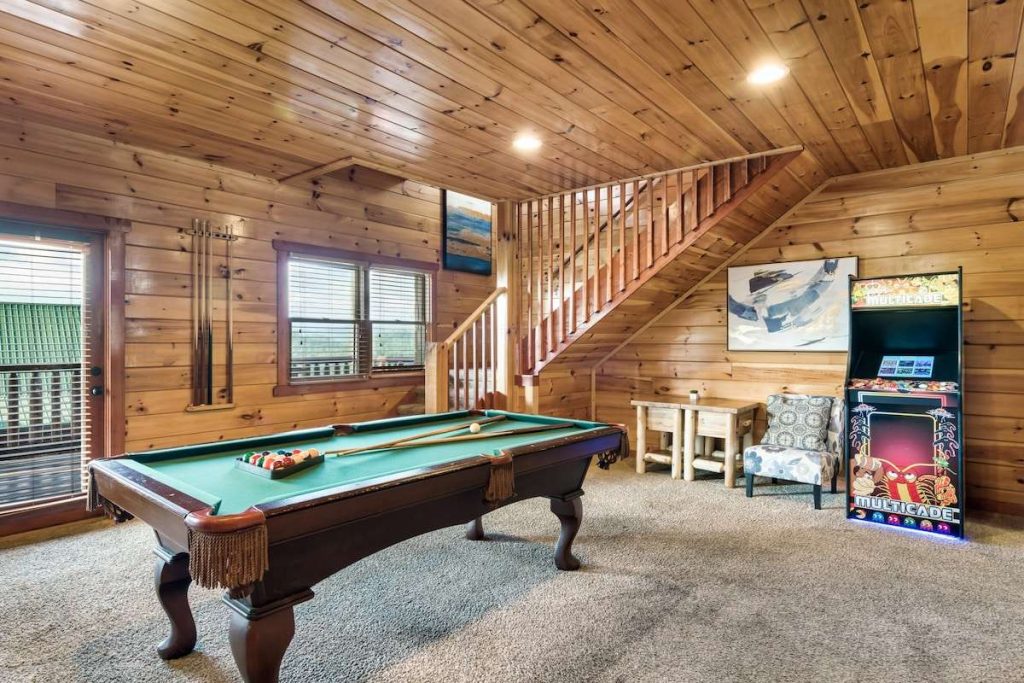 A rustic game room with wood-paneled walls and ceilings. In the center is a classic pool table with green felt and billiard balls. To the right, a vintage-style Multicade arcade machine stands next to a small wooden table and a patterned armchair. A wooden staircase with slats is visible in the background.