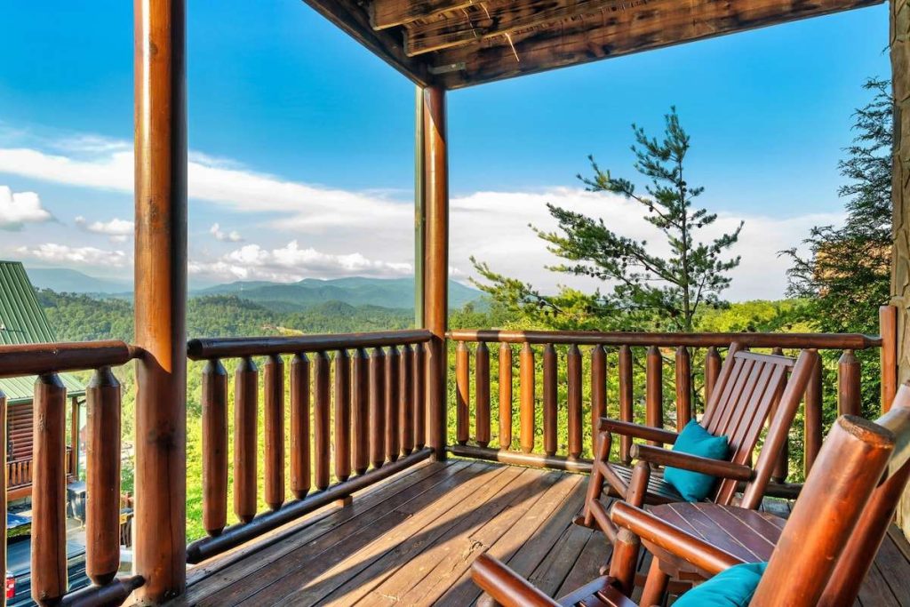 A rustic wooden balcony with log railings and rocking chairs overlooking a vast, lush mountain range under a blue sky.