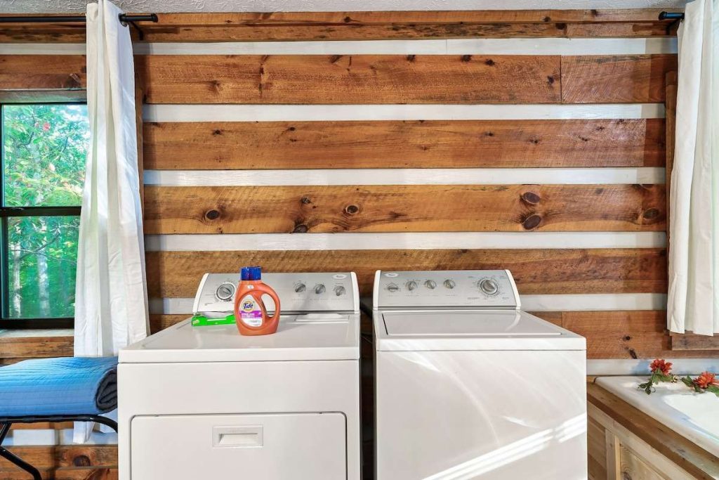A laundry area with a white top-load washer and dryer set against a rustic wood-and-white chinked log wall, featuring a bottle of detergent and a nearby window.