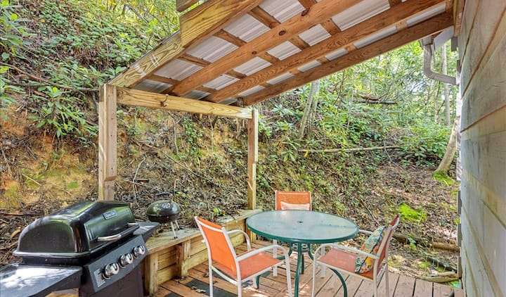 A rustic outdoor patio area nestled against a wooded hillside. The wooden deck is covered by a simple slanted corrugated metal roof supported by timber beams. The space features a black gas grill, a small charcoal kettle grill, and a green circular table with three orange mesh chairs.