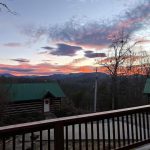 A vibrant orange and purple sunset over a mountain range, viewed from a wooden balcony overlooking log cabins with green metal roofs.
