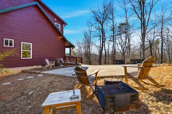 A backyard area with a concrete pad, wooden rocking chairs, and a charcoal park-style grill next to a red cabin.