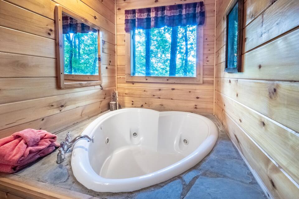 A white, heart-shaped jetted tub built into a stone surround within a rustic wooden cabin room, featuring a window overlooking a lush green forest.