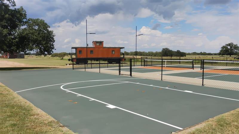 An outdoor sports complex featuring a green basketball half-court in the foreground and fenced-in pickleball courts behind it, with a vintage orange train caboose parked in the background under a cloudy sky.